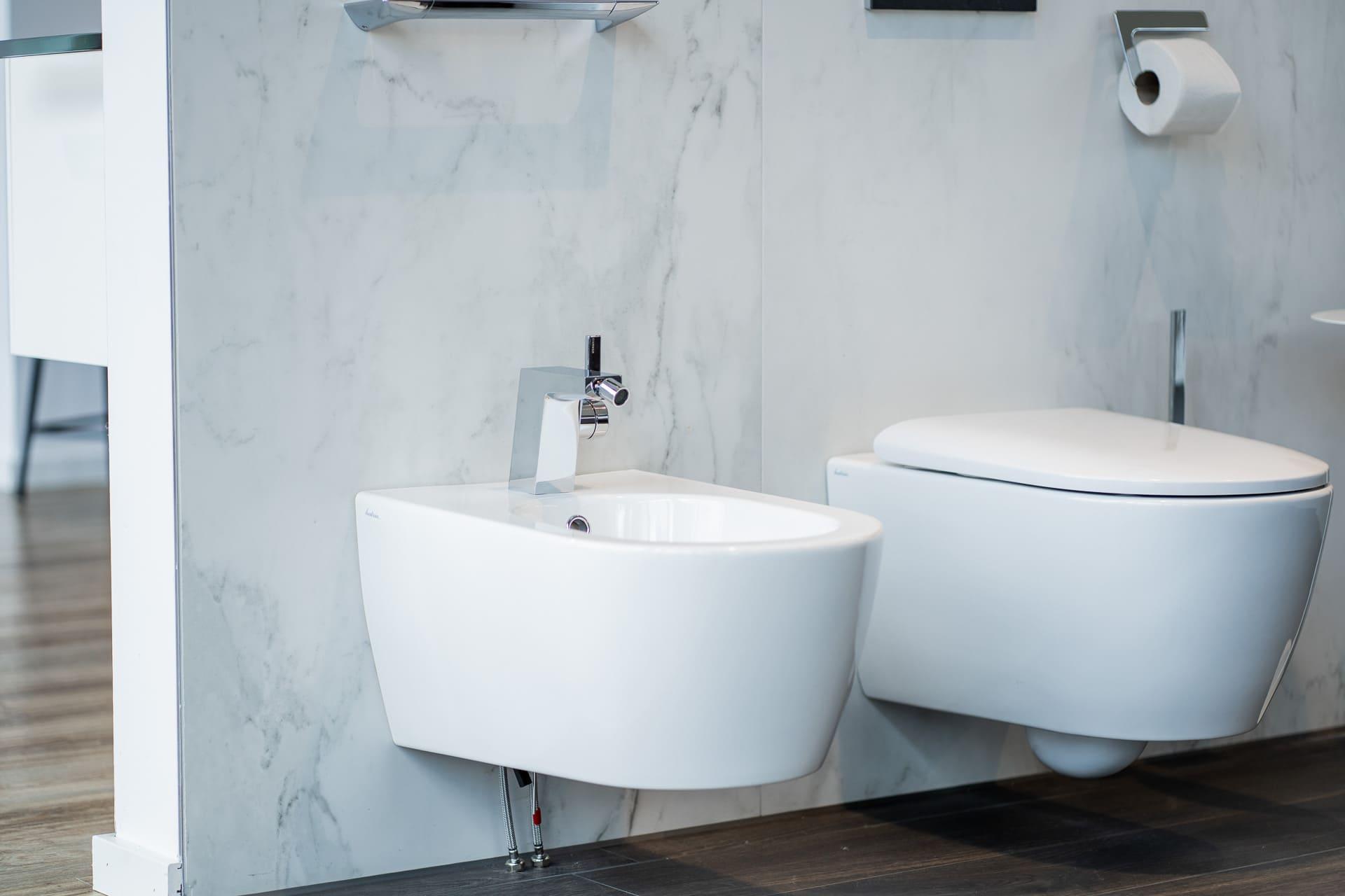 Ein modernes Bidet in Wandmontage mit edler Marmoroptik, daneben ein weißes Wand-WC. Rechts befindet sich ein eleganter silberner Toilettenpapierhalter, der funktionale und stilvolle Akzente setzt.