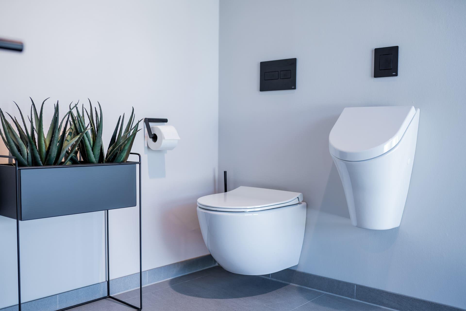 Ein stilvolles Badezimmer mit einem weißen Wand-WC und einem Urinal, beide ausgestattet mit einer eleganten schwarzen Betätigungsplatte. Auf der linken Seite befindet sich eine moderne Pflanze in einem Blumenständer, und ein praktischer Toilettenpapierhalter ist an der Wand montiert.